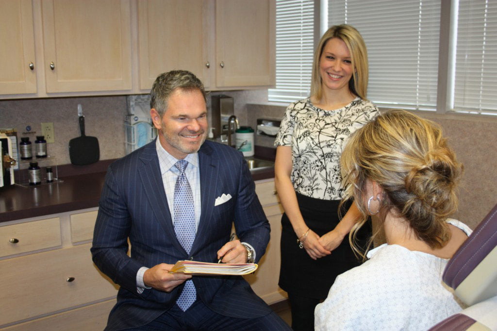 doctor holding folder listens to and smiles at patient while assistant stands in background